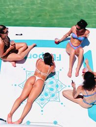 Aerial view of four people in colorful bikinis lounging on a large white-and-blue floating mat with a mandala design, holding drinks while drifting on clear turquoise water