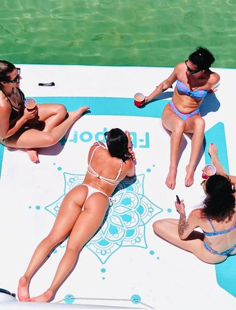Aerial view of four people in colorful bikinis lounging on a large white-and-blue floating mat with a mandala design, holding drinks while drifting on clear turquoise water