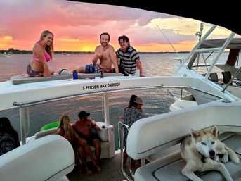 Group of people relaxing on a boat at a vibrant pink-orange sunset over a lake — three people lounging on the upper deck and a husky sprawled on a cushioned seat with boats on the water.