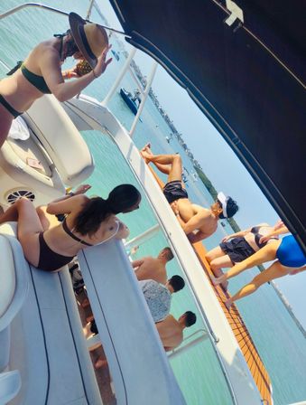 Group of people in swimwear lounging on a sunny yacht over turquoise coastal bay, one person with a sunhat holding a pineapple drink, others relaxing on the deck with boats and shoreline visible in the distance.