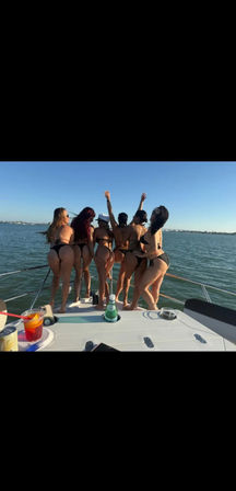 Six women in black bikinis posing at the bow of a boat, backs to camera with arms raised, overlooking a calm blue coastal bay and distant shoreline on a sunny day with drinks on deck.