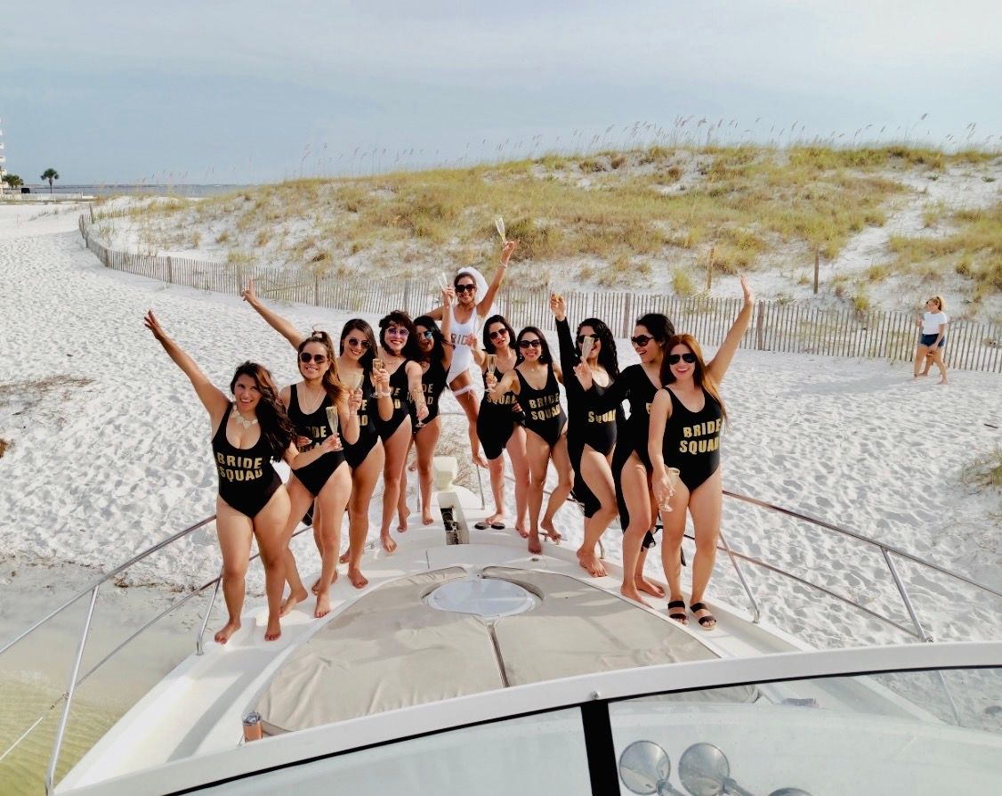 Bachelorette party on a boat at a sandy beach — women in matching black "BRIDE SQUAD" swimsuits and one in white sash cheering with champagne on the bow in front of coastal dunes.