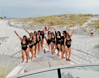 Bachelorette party on a boat at a sandy beach — women in matching black "BRIDE SQUAD" swimsuits and one in white sash cheering with champagne on the bow in front of coastal dunes.