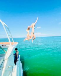 Two people in bikinis mid-air jumping off a yacht into turquoise ocean on a sunny summer day, another person leaning on the boat rail watching.