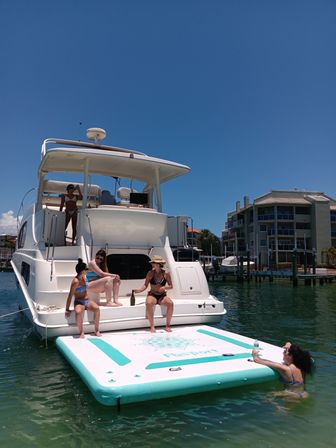 Group of friends relaxing on the stern of a white yacht at a sunny marina, lounging on a turquoise inflatable dock near waterfront condos.