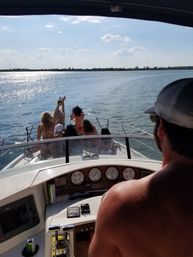 Group of people cheering on the bow of a motorboat cruising a sunlit lake, captain at the helm, sparkling water and tree-lined shoreline under a blue sky