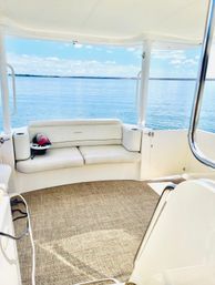 Sunny boat deck with white cushioned bench, woven marine carpet and a cap on the seat, overlooking calm blue lake and clear sky horizon.