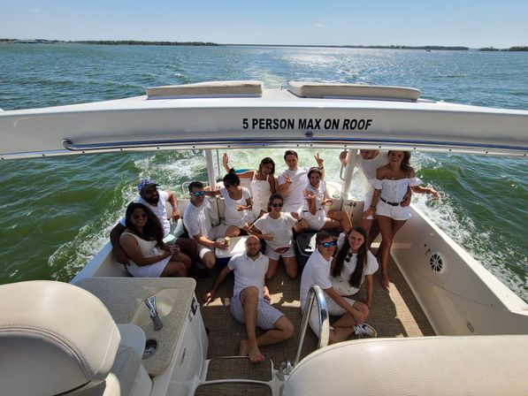 Group of friends in white outfits enjoying a sunny boat party on open water, wake trailing behind the motorboat and a '5 PERSON MAX ON ROOF' sign visible.