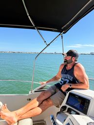 Man with beard and cap relaxing at the helm of a motorboat under a black bimini, legs outstretched over the bow, cruising calm turquoise coastal bay with a distant shoreline and clear blue sky.