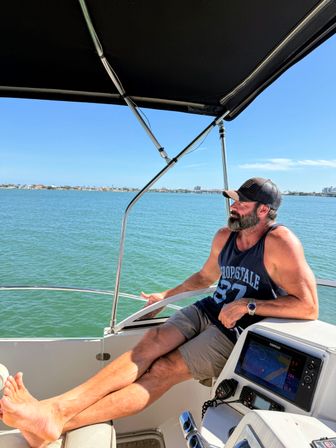 Man with beard and cap relaxing at the helm of a motorboat under a black bimini, legs outstretched over the bow, cruising calm turquoise coastal bay with a distant shoreline and clear blue sky.