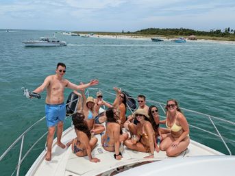 Boat party on a sunny summer day — a group of friends in swimsuits on a yacht bow in turquoise coastal waters, with anchored boats and a sandy beach island visible in the background.