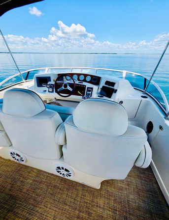 White boat helm with twin cushioned captain chairs, wooden steering wheel and instrument panel overlooking calm blue lake and sunny sky — ready for a relaxing cruise