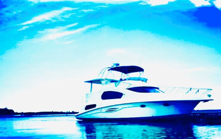 Sleek white motor yacht anchored on calm coastal waters with a bright blue sky and shoreline reflected in the glassy water.
