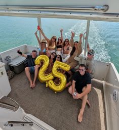 Smiling friends on a boat waving and holding gold "25" balloons beside turquoise open water and a foamy wake during a sunny summer boat party.