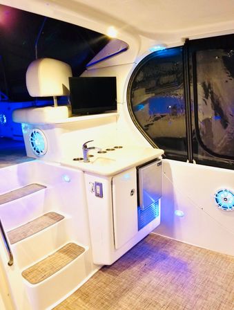 White yacht cabin galley and boat interior featuring a sink with faucet, built-in mini fridge, flat-screen TV, carpeted steps, speakers and blue LED accent lighting