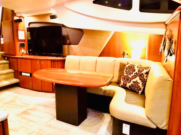 Cozy yacht salon dining area with curved beige leather banquette, glossy wood table, patterned accent pillow, flat-screen TV and warm ambient lighting