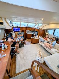 Group of friends relaxing onboard a luxury yacht — wood-paneled salon with large windows, kitchenette, drinks on the table and a curious husky dog.