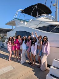 Group of women in pink and white outfits smiling and posing on a dock beside a large white yacht at a sunny marina with clear blue sky