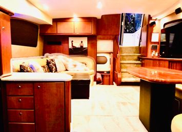 Cozy luxury yacht salon with curved cream leather sofa, patterned pillows and faux fur throw, glossy wood cabinetry, built-in entertainment, carpeted floor, dining table and carpeted stairs leading up to the cockpit