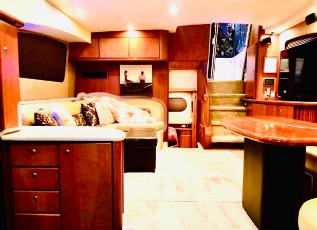Cozy luxury yacht salon with curved cream leather sofa, patterned pillows and faux fur throw, glossy wood cabinetry, built-in entertainment, carpeted floor, dining table and carpeted stairs leading up to the cockpit