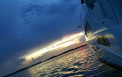 Sleek white yacht hull reflecting a dramatic blue-and-orange sunset over rippling coastal waters with a low silhouetted shoreline and moody clouds.