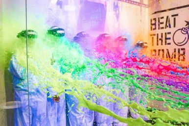 Group of people in white protective suits and helmets inside an indoor paint-splash room, bright neon green, pink and purple paint exploding mid-air across a glass wall.