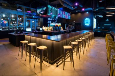 Neon-lit modern bar interior with a long wood-topped island, rows of metal stools, digital menu screens and beer taps under purple-blue industrial lighting