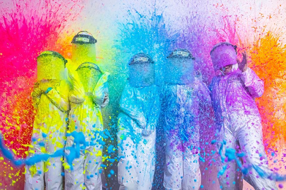 Group wearing white coveralls and helmets blasted with buckets of rainbow paint—vivid yellow, blue, purple and pink splashes at a lively color festival paint explosion