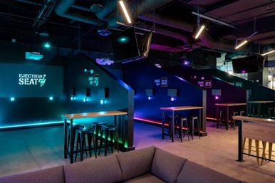 Modern esports lounge with numbered private gaming booths, neon blue-purple lighting, high-top tables and stools, and sofa seating.