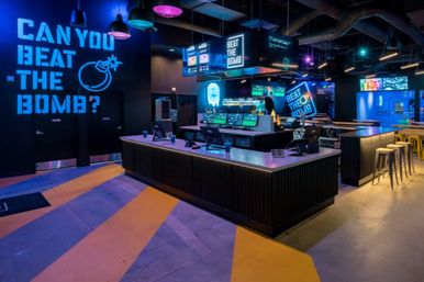 Neon-lit indoor arcade-bar counter with touchscreen registers and stools, colorful purple and blue lighting and large 'Can you beat the bomb?' wall signage.