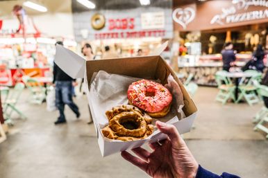 Hand holding a takeaway box with a pink sprinkle donut and a brown-glazed cruller in a lively indoor food market with blurred tables and shoppers