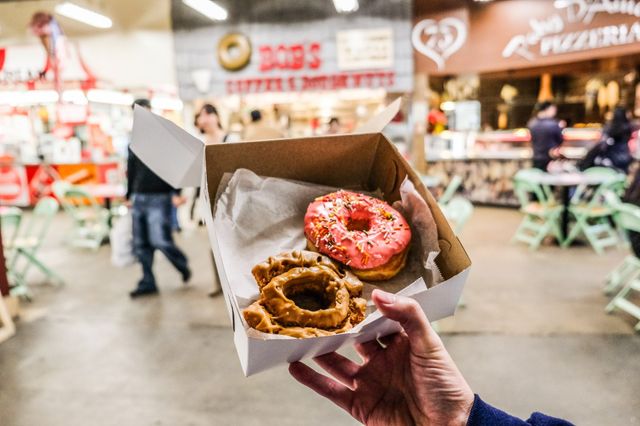 Hand holding a takeaway box with a pink sprinkle donut and a brown-glazed cruller in a lively indoor food market with blurred tables and shoppers