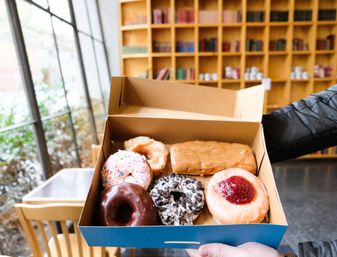 Assorted donuts (chocolate, sprinkle, cookies-and-cream, jelly) in a blue box held inside a cozy café with large windows and wooden bookshelves
