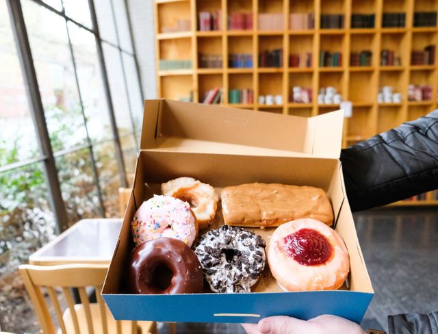 Assorted donuts (chocolate, sprinkle, cookies-and-cream, jelly) in a blue box held inside a cozy café with large windows and wooden bookshelves