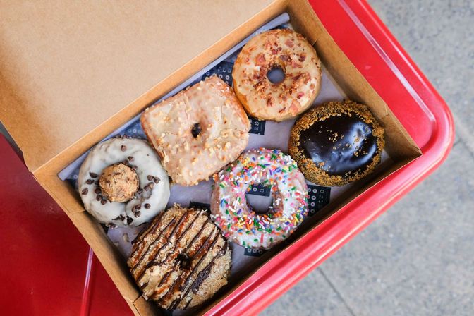 Top-down view of assorted artisan donuts in a cardboard takeout box on a red tray — colorful sprinkle, chocolate-glazed, maple, square-glazed and cookie-topped varieties.