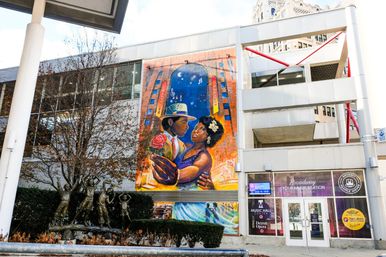 Vibrant street mural of a romantic dancing couple under a starry arch on a downtown building facade, beside a modern white entrance and a bronze children's sculpture in a streetside plaza.