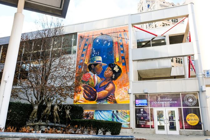 Vibrant street mural of a romantic dancing couple under a starry arch on a downtown building facade, beside a modern white entrance and a bronze children's sculpture in a streetside plaza.