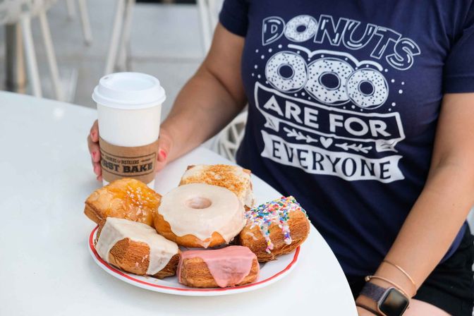 Plate of assorted glazed, frosted, and sprinkled donuts next to a takeaway coffee cup on a cafe table, person wearing a 'Donuts are for everyone' shirt.