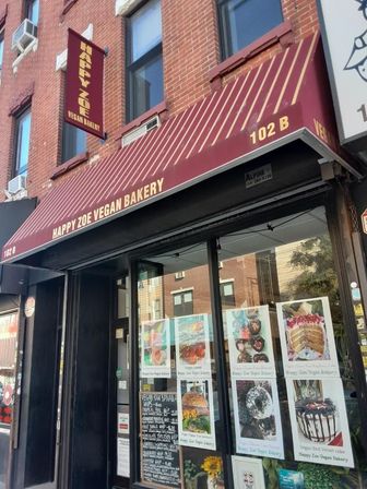 Maroon striped awning over a red brick vegan bakery storefront, glass window filled with colorful photos of cakes, pastries and sandwiches.