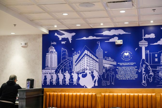 Bakery cafe interior with a bold blue city-skyline mural featuring an airplane, dancing and walking silhouettes above tan booth seating and a person at the service counter