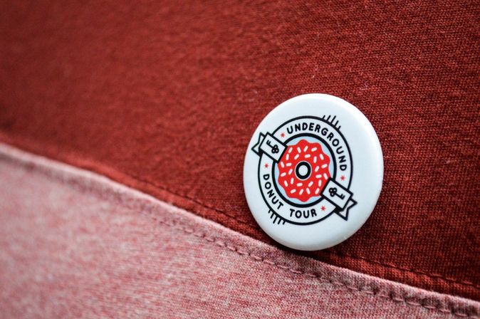 Close-up of a white pin-back button with a playful red sprinkled donut logo pinned to layered red and pink fabric