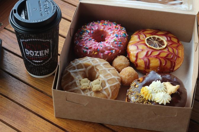 Assorted colorful gourmet donuts in a cardboard box — pink sprinkle, citrus-glazed with dried slice, maple with mini cookies and chocolate-topped with cream — next to a takeaway coffee cup on a wooden cafe table.