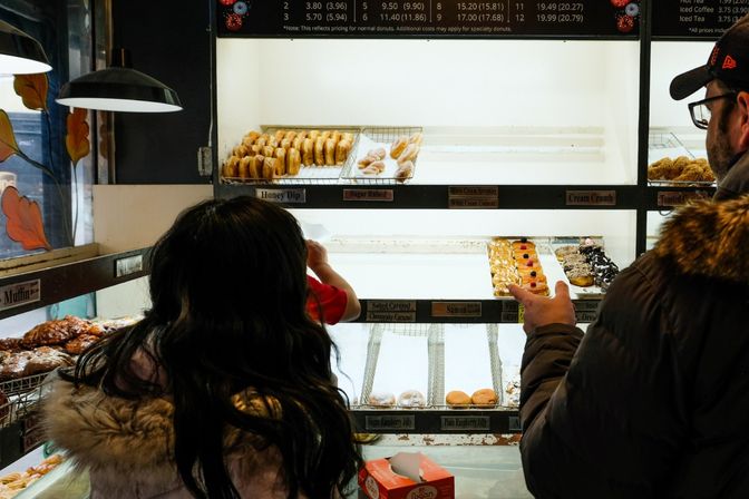 Customers in winter coats choose assorted glazed, filled, and sprinkle-topped donuts from a brightly lit donut shop display case.