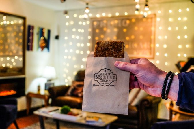 Hand holding a spiced shortbread cookie in a small kraft paper bag with a stamped logo, cozy living room backdrop with warm string lights and a glowing fireplace