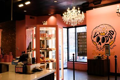 Bright pink donut shop interior with a glass display of colorful donuts, a sparkling crystal chandelier, and a playful skull-shaped donut mural by large front windows