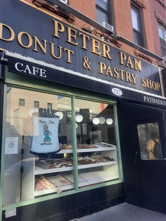Urban donut and pastry shop storefront with black-and-gold sign, green-framed window showcasing trays of assorted donuts and pastries and warm cafe globe lights inside.