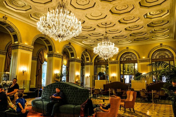 Grand luxury hotel lobby with crystal chandeliers, ornate coffered ceiling, arched windows and velvet seating, guests relaxing.