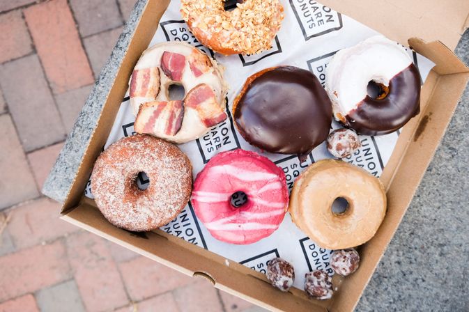 Assorted gourmet donuts in a cardboard box on a brick city sidewalk — bacon-topped, chocolate-glazed, pink-frosted, cinnamon-sugar, maple glaze and donut holes.