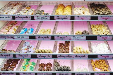 Bright pink-lined bakery display case filled with assorted colorful donuts and pastries — sprinkles, chocolate crullers, cronuts and croissants.