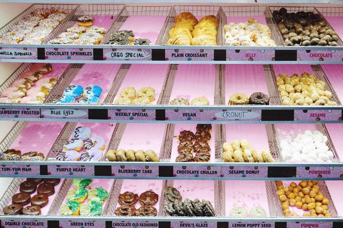 Bright pink-lined bakery display case filled with assorted colorful donuts and pastries — sprinkles, chocolate crullers, cronuts and croissants.